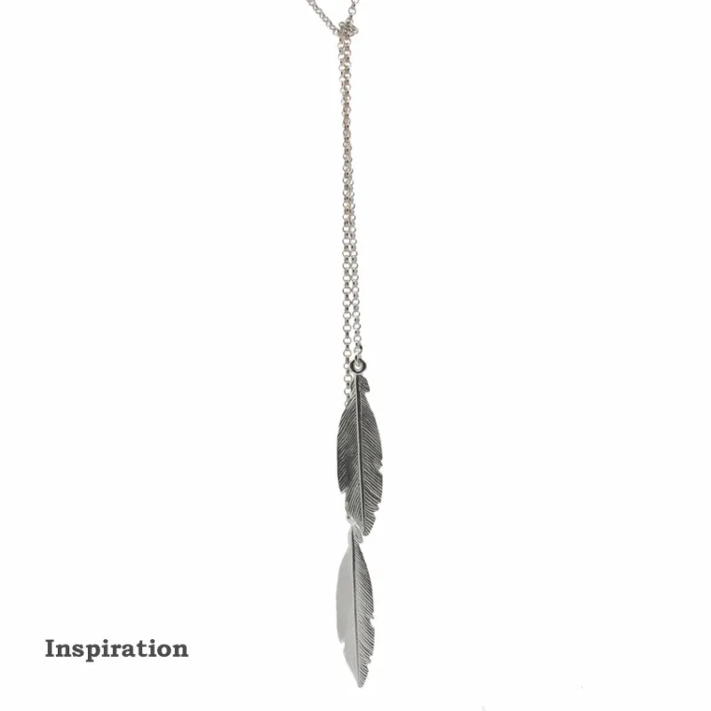 Ett Silverkedja ärtlänk YD 1,8mm - 10cm halsband har två fjäderhängen i olika längder. "Inspiration" visas i fet svart text nere till vänster på en vit bakgrund.