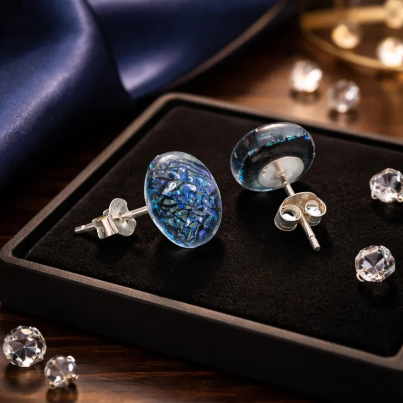 Ett par Örstift med platta 5mm i Sterlingsilver vilar på en smyckesbricka i svart sammet, omgiven av lösa klara ädelstenar, med mörkblått satintyg delvis synligt i bakgrunden.