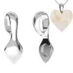 Två Spetsögla 16 mm sterling silver (limmas) skedformade hängen visas från olika vinklar, bredvid ett halsband med ett krämigt, hjärtformat hänge på en silverkedja. Bakgrunden är vit.