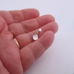 En hand med guldring på ringfingret håller i Spetsögla 16mm sterlingsilver - limmas, ett litet runt silverhänge med bygel, mot en slät vit bakgrund.