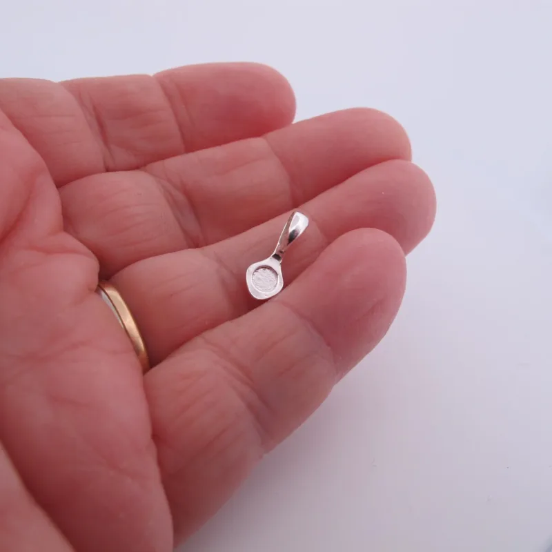 En hand med guldring på ringfingret håller i Spetsögla 16mm sterlingsilver - limmas, ett litet runt silverhänge med bygel, mot en slät vit bakgrund.