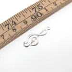 En Silverberlock Musiktema - G-Klav berlock vilar på en vit yta bredvid en trälinjal som visar att den är ca 2,5 cm lång.