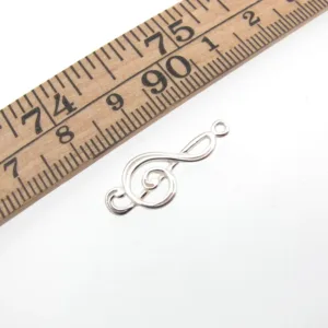 En Silverberlock Musiktema - G-Klav berlock vilar på en vit yta bredvid en trälinjal som visar att den är ca 2,5 cm lång.