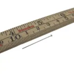 En Hattpinnar 25-50mm/0,51 metallnål i sterlingsilver är placerad bredvid en centimetermarkerad linjal i trä märkt "Sweden" i rött, vilket indikerar att nålens längd är cirka 3 centimeter.