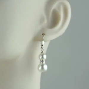 Närbild av ett skyltdocksöra med ett dinglande örhänge bestående av två vita pärlor åtskilda av en Hattpinnar sterlingsilver 25-50mm/0,51 på en krok. Bakgrunden är enfärgad och ljus.