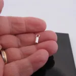 En hand håller i en Cylinderformad avslut för rem 3mm, ett litet silverfärgat cylindriskt avslut med ögla, mot ett svart rektangulärt objekt och vit bakgrund.