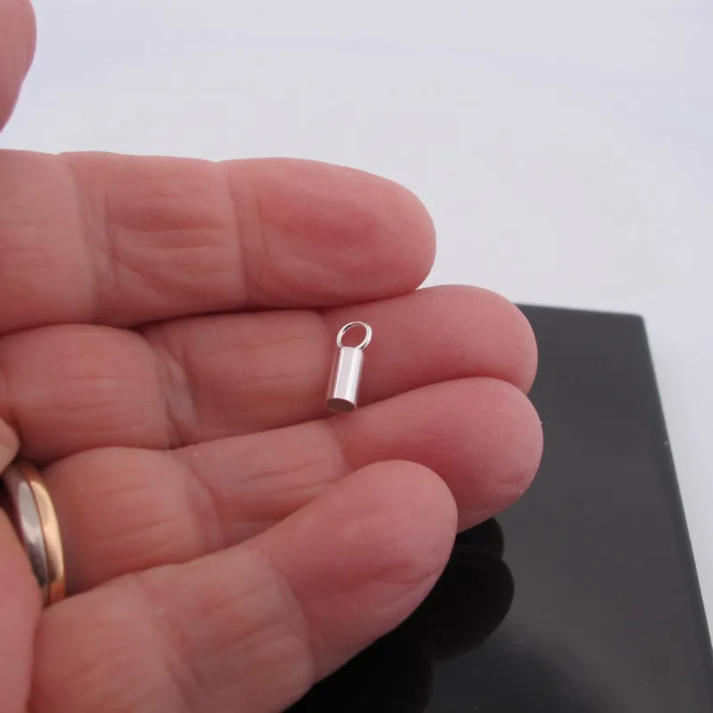 En hand håller i en Cylinderformad avslut för rem 3mm, ett litet silverfärgat cylindriskt avslut med ögla, mot ett svart rektangulärt objekt och vit bakgrund.