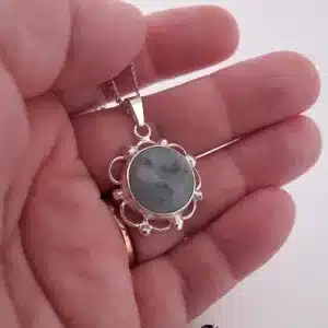 En hand håller i "Halsband med handslipad Kalcedon 32x23 mm", ett silverhalsband med en rund, ljusblå kalcedon infattad i en utsmyckad infattning, mot en vit bakgrund.
