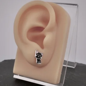 Örhänge Emaljerad Svart Katt visas på en realistisk, hudfärgad modeleöron, monterad på ett genomskinligt akrylställ mot en mörk bakgrund.