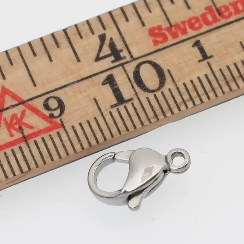 A Karbinhake kirurgiskt stål 13mm ligger bredvid en trälinjal och visar sin längd på cirka 1,3 centimeter.