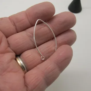 En hand håller i en Öronkrok i Sterlingsilver Marquises, ett fint, droppformat örhänge i sterlingsilvertråd med en liten ögla i botten; personen bär en vigselring i guld och en annan ring.