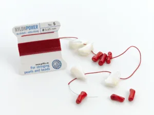 En rulle Smycketråd Griffin Nylon Power Svart med vita snäckpärlor och röda korallpärlor som delvis är uppträdda på den svarta nylontråden, plus några lösa pärlor, mot en vit bakgrund.