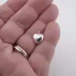 En hand med en ring i guld och silver håller Berlock sterlingsilver - Litet Hjärta, ett litet, blankt, hjärtformat hängsmycke i sterlingsilver, mot en vit bakgrund.