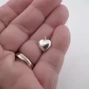 En hand med en ring i guld och silver håller Berlock sterlingsilver - Litet Hjärta, ett litet, blankt, hjärtformat hängsmycke i sterlingsilver, mot en vit bakgrund.