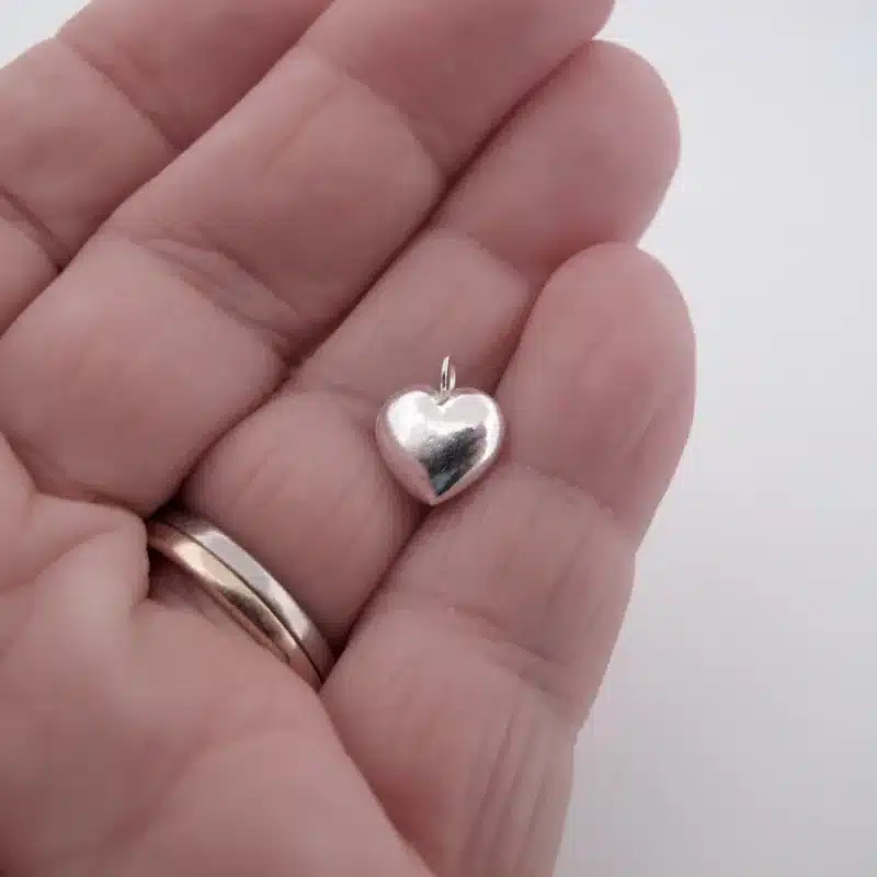 En hand med en ring i guld och silver håller Berlock sterlingsilver - Litet Hjärta, ett litet, blankt, hjärtformat hängsmycke i sterlingsilver, mot en vit bakgrund.