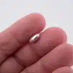 Närbild av en hand som håller i Berlock sterlingsilver - Litet Hjärta, en liten, glänsande, hjärtformad berlock i sterlingsilver med ögla på toppen, mot en vit bakgrund.