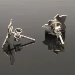 Ett par Örstift med platta 5mm i Sterlingsilver visas på en blank svart yta, med ett örhänge liggande platt och det andra upprätt, och visar både fram- och baksidan tillsammans med sina stolpar.