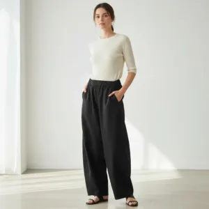 I Bomullsbyxa - Mood står en kvinna inomhus i lösa svarta byxor med händerna i fickorna, tillsammans med en vit långärmad topp och sandaler. Naturligt ljus flödar in genom stora fönster när hon tittar framåt.