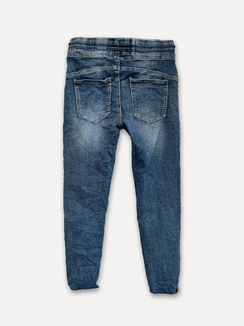 The Cozy elastisk byxa - Jeans är blå denimbyxor, visade bakifrån med två fickor och en elastisk linning, liggande platt på en vit bakgrund.
