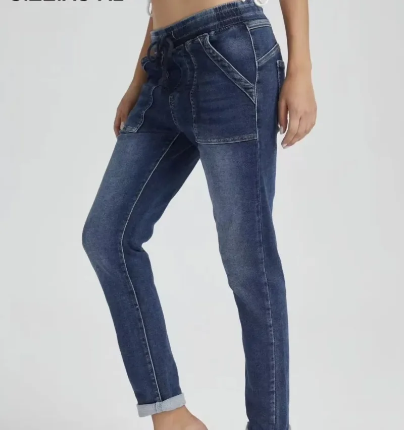 En person modellerar Jogger Jeans elastisk byxa-högmidjiga blå jeans med framfickor och dragsko i midjan-stående med ett ben lätt böjt mot en ljus bakgrund.