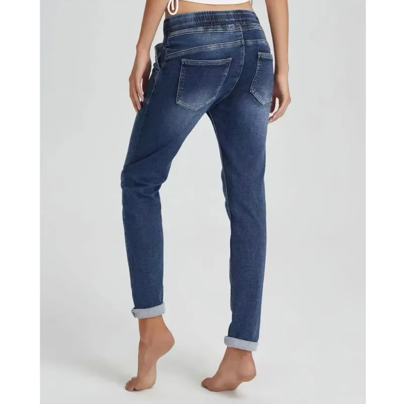 En person står barfota mot en ljus bakgrund, sedd bakifrån, iklädd Jogger Jeans elastisk byxa-blå jeans med upprullade ben, bakfickor och resår i midjan.