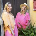 Två blonda kvinnor med pastellrosa och persikofärgade Linneblus - ELBA linnetoppar står utanför en byggnad med brun dörr och gul vägg och ler. En av dem bär hatt och i förgrunden syns en grön ormbunke.