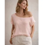 En kvinna med vågigt blont hår bär Bomullstopp - Soleil flera färger i ljusrosa och ljusa byxor, står mot en neutral bakgrund och tittar åt sidan med ett avslappnat uttryck.