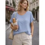 En kvinna står på en stadsgata kantad av byggnader och kaféer, håller i en baguette och bär Bomullstopp - Soleil flera färger blus i ljusblått med beige byxor, kort blont hår och en beige axelremsväska.