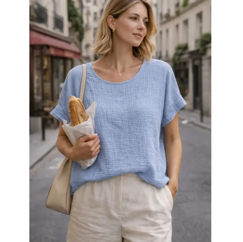 En kvinna står på en stadsgata kantad av byggnader och kaféer, håller i en baguette och bär Bomullstopp - Soleil flera färger blus i ljusblått med beige byxor, kort blont hår och en beige axelremsväska.