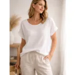 Iförd Bomullstopp - Soleil flera färger står en kvinna med vågigt blont hår och beigea byxor med dragsko inomhus med ena handen i fickan, tittar åt sidan och ler mjukt.