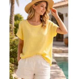 En kvinna står vid en pool utomhus, ler och tittar åt sidan, klädd i Bomullstopp - Soleil flera färger i gult med vita shorts och en stråhatt. I bakgrunden syns gröna växter och en byggnad.