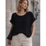 En kvinna står utomhus klädd i Bomullstopp - Soleil flera färger, beige byxor och en svart axelremsväska. Hon har axellångt blont hår och tittar åt sidan i en gatumiljö i en stad.