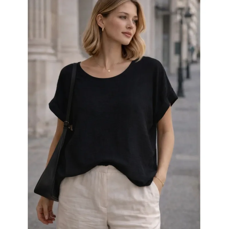 En kvinna står utomhus klädd i Bomullstopp - Soleil flera färger, beige byxor och en svart axelremsväska. Hon har axellångt blont hår och tittar åt sidan i en gatumiljö i en stad.