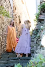 Två kvinnor som bär Linneklänning Sicily underbar sommarklänning går uppför en stentrappa mellan rustika väggar och grönska i starkt solljus, ler och njuter av en charmig, fridfull utomhusmiljö.