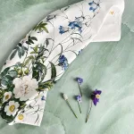 White scarf with a blue and green botanical print laid on a pale green textured surface; a few small flowers lie nearby for styling.