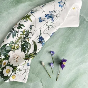White scarf with a blue and green botanical print laid on a pale green textured surface; a few small flowers lie nearby for styling.
