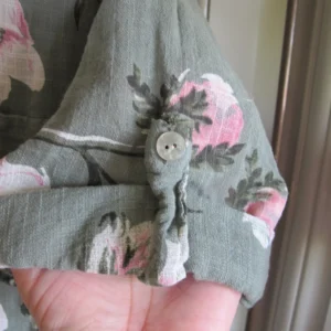 Close-up of a green floral garment with pink blossoms and a white button; a hand is visible at the bottom edge.