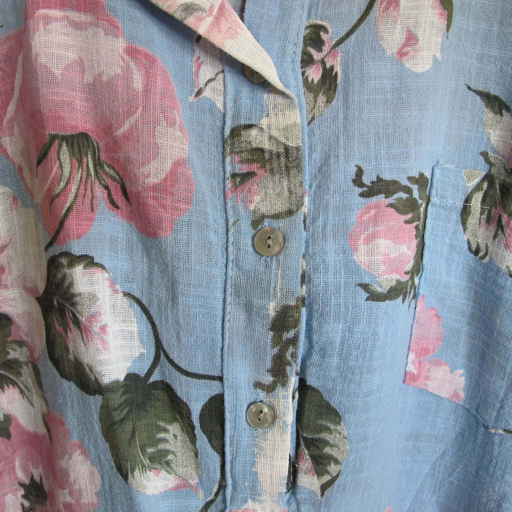 Annie Ljust Blå Blommig Bomullsskjorta 4 Close-up of blue floral shirt fabric with pink roses, green leaves, and a button placket visible in the center area.