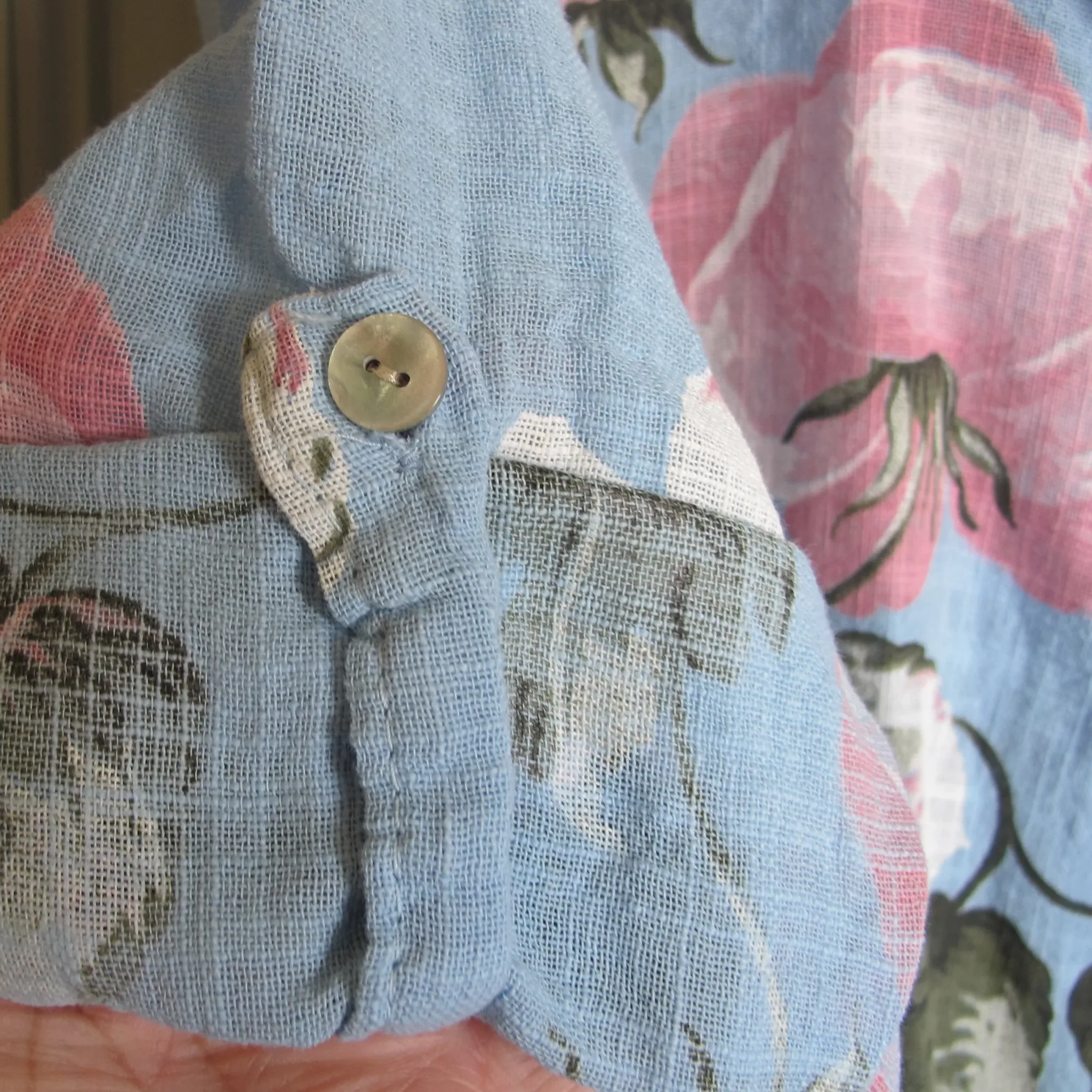 Annie Ljust Blå Blommig Bomullsskjorta 5 Close-up of light blue denim fabric with a round metal button, with a pink floral textile in the blurred background.