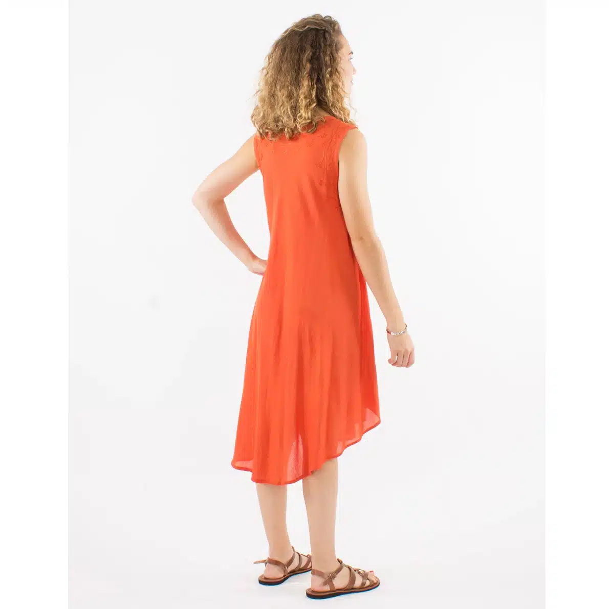 Woman in an orange sleeveless midi dress, seen from the back, standing with one hand on hip and wearing strappy sandals.
