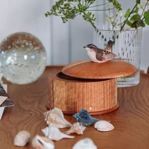 Wooden round box with its lid slightly open, a small bird figurine perched on top, surrounded by seashells on a wooden table.