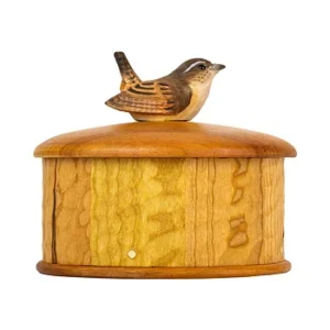 Wooden cylindrical birdhouse with a small carved bird perched on the lid on natural-toned wood.