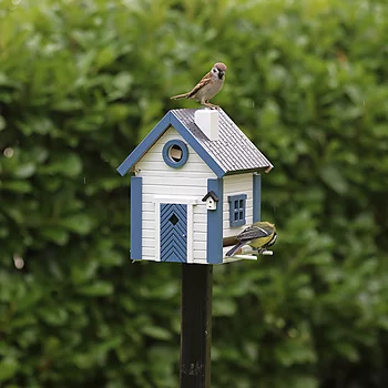 Small blue-and-white birdhouse on a black post with two sparrows perched on it, green leafy background.